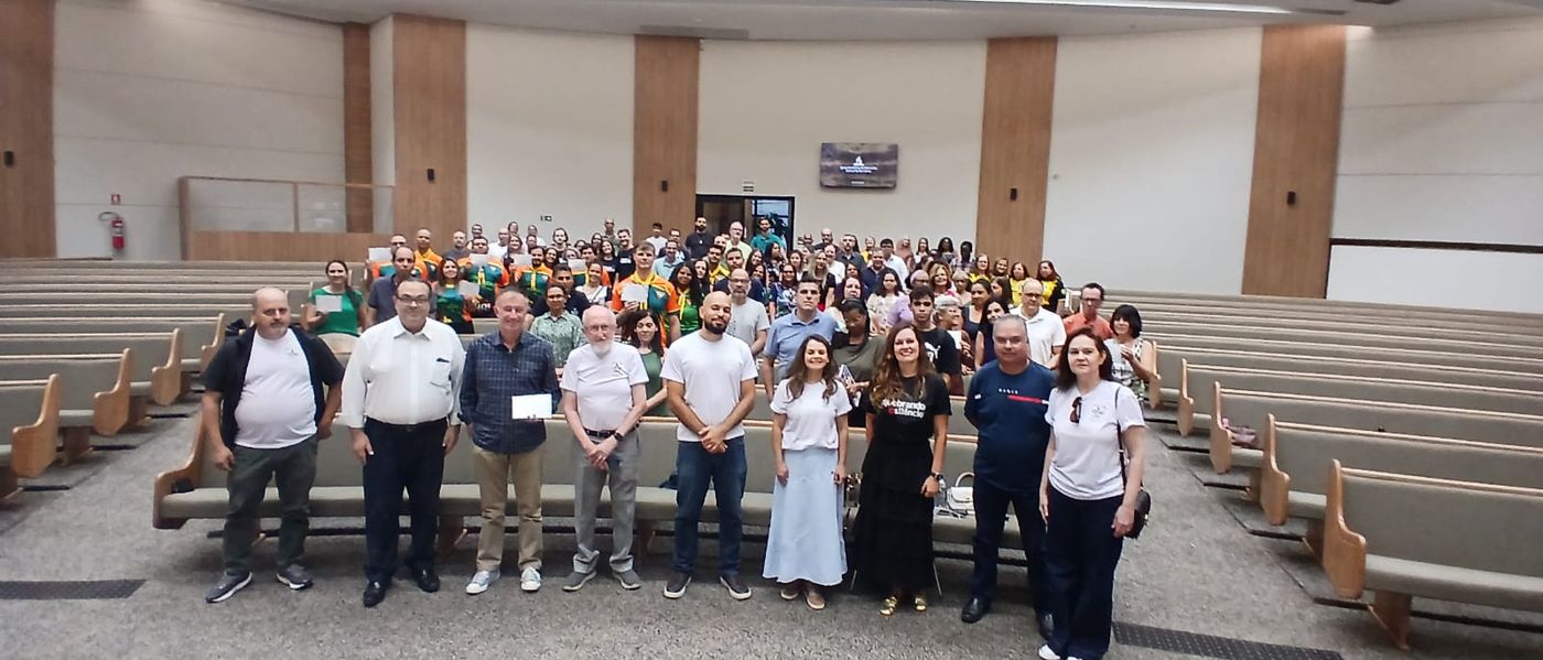 Vista ampla do auditório cheio, com pessoas de todas as idades nos bancos e a equipe reunida à frente